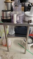 View of stainless steel two-shelf kitchen shelf with various kitchen equipment on the top shelf.