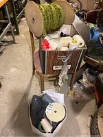 Wide view showing a large wooden spool with green ornamental fabric trim on top of a chair and a box with various sewing supplies including zippers and smaller spools.