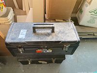 Photo of two stacked Black & Decker plastic tool boxes with metal diamond plate lids, placed indoors on a concrete floor near cardboard boxes and other items
