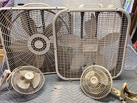 Four fans include two large square Lasko box fans and two small clamp-on fans displayed on a quilted surface, showing condition and size.