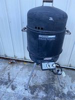 Front view of Brinkmann Smoke 'N Grill outdoor gas cooker showing black cylindrical main body with lid closed, mounted on three tripod metal legs, with attached propane regulator and control knobs