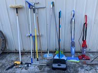 Photo showing an assortment of window and floor cleaning tools including mop heads, squeegees, brooms, and lightweight cordless vacuums, arranged against a metal wall.