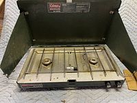 Top view of Coleman propane camping stove showing two burners, metal grate, and control knobs. Stove has side wind guards and some signs of use.
