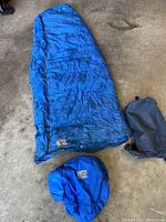 Blue down sleeping bag fully laid out on concrete surface with matching hood and a dark stuff sack next to it