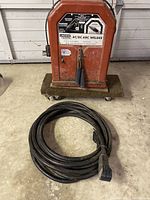Red Lincoln Electric AC/DC Arc Welder unit on a wheeled dolly with heavy duty power cord neatly coiled in front.