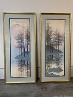 Two vertical landscape prints featuring trees reflected in water, framed in gold with glass.