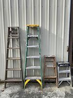 Front view of four ladders leaning against siding: 6ft wood, 6ft fiberglass green, 3ft wood, and 2ft aluminum with black top