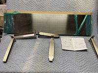 Three rectangular stainless steel look shelves wrapped in green plastic tape, accompanied by four stainless steel mounting brackets and an assembly instruction sheet laid out on a padded surface.