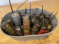 Top-down view of 28 vintage oil cans clustered inside a galvanized metal tub, showing various sizes, rust, and dirt on cans.