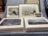 Two stacks of matted photos and four black frames with maritime and landscape themes arranged on a quilted blanket