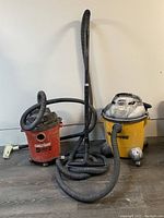 Photo showing both shop vac units, yellow Shop-Vac and red Genius vac with hoses and attachments, placed on floor near wall.