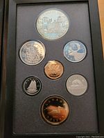 Photo showing full set of seven coins in a black display case including the large commemorative silver dollar coin and six smaller coins. Case shows some damage.