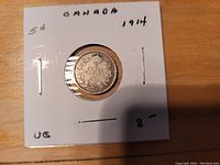 Front side of 1914 Canadian Silver 5 cent coin secured in a white paper holder with handwritten markings related to coin details and price.
