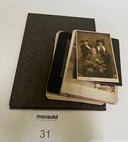 Stacked vintage black and white photographs with black and brown mount boards alongside a black album or book.