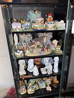 Full view of black display cabinet showing four shelves filled with various figurines: nativity sets in white ceramic and colorful sets, Disney Mickey and Minnie figurines, Christmas village buildings and snow globes.