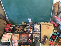 Photo showing the entire lot of board games, computer games, checkers, cards, bingo pieces, chalkboard, and toy trucks arranged in front of a green chalkboard