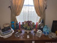 Wide view of all items on a wooden table beneath window with curtains. Shows Capodimonte floral centerpiece, candle holders, plastic containers, glass vase, and other items.
