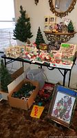 Photo showing overall collection of vintage Christmas decorations including village buildings, fiber optic trees, boxes of Christmas lights and ornaments, decorative plate and figurines.