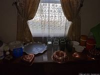 Full view of all copper items, glasses, Thermos, kitchenware, and decorative pieces on table in front of window
