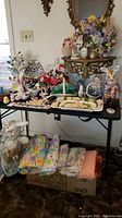 Overall view of table filled with various Easter decorations including bunnies, eggs, baskets, and floral displays, as well as folded pastel linens underneath the table on a box.