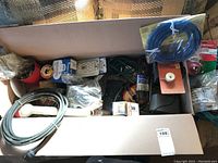 Wide view of box containing assorted electrical and plumbing repair supplies including wires, cords, hoses, and multiple small packaged items.