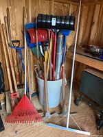 Tools including shovels, rakes, hoes, brooms arranged in a bucket and against a wall inside a wooden shed.