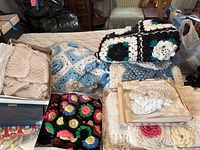 Overview showing multiple folded hand knitted afghans in black and white, blue and white, multicolor flower patterns, and cream colors, along with boxes containing baby clothes such as sweater sets and booties.