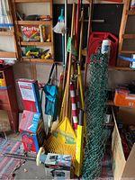 Photo showing multiple garden rakes, brooms, green metal fencing rolls, two American flags with poles, some tool boxes, and other miscellaneous outdoor tools.