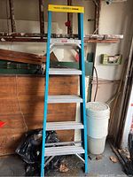 Photo shows a blue metal step ladder with a yellow top cap, positioned upright in a garage-like setting with tools and storage in the background.