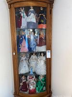 Full wooden curio cabinet holding the 12 Musical Calendar Miss dolls arranged in four rows of three dolls each, showing a variety of period-style dresses representing months from January to December.