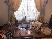 View of the entire lot placed on a wooden table showing cheese tray with glass cover, two German beer steins with lids, carousel horse figurine, a shot glass figurine, music box, vintage cookbooks, wicker paper plate holders, and white plastic trays