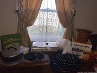 Photo showing various cookware and textiles including Crock-Pot box, ceramic griddle, metal covered dish, placemats, and white tablecloth on wooden table