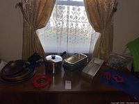 Photo of various kitchen pans including frying pans, stainless steel pot with lid, and meatloaf/bread pans on a wooden table near a window with curtain backdrop.