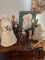 Various religious ceramic statues and decor including Virgin Mary, nuns, girl with flowers, Pieta statue, and religious relief plaque on wood dresser