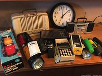 Photo of vintage radios including cream-colored and smaller black radio, white wall clock, six flashlights (one boxed), and a white calculator.