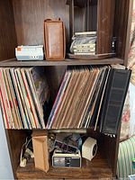 Shelving unit with multiple vinyl records arranged vertically, cassette player and cassettes on upper shelf, and two vintage alarm clocks along with other small items on lower shelf.