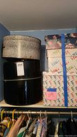 Photo showing black and white decorative round hat box stacked on black Hess carrying case with textured faux reptile finish. Partial view of multiple Hess branded boxes and floral patterned box to the right. Clothing hangers below.