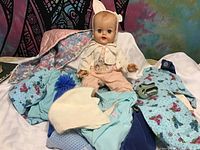Vintage baby doll dressed in handmade clothing with bonnet and additional clothing and shoes surrounding it