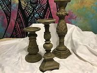 Three metal antique style candlesticks arranged from shortest to tallest against a cloth backdrop and colorful tapestry.