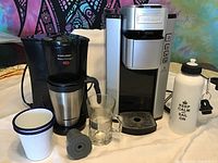 Photo showing Cuisinart coffee maker, Black & Decker Brew N Go coffee maker with travel mug, two white enamel mugs, clear glass mug and water bottle.