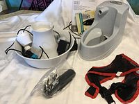 Two pet water fountains (one ceramic white and one gray plastic), pet trimmer, and pet harness laid out on a cloth surface.