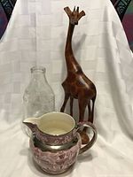 Photo showing all three items: the Wedgwood Ferrara pitcher in front, clear glass milk bottle on left, and wooden hand-carved giraffe on right.
