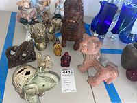 Overview of the eight Asian decor pieces, showing a mix of metal and porcelain figurines including a brown metal lion, golden dragon figure, small painted porcelain beings, and two animal statues in front: a ceramic elephant and a reddish dog figurine with fur tail.