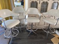Four white leather bar-height chairs arranged in a row showing front and side views. Chairs have padded seats and backs with chrome armrests and bases. Some scratches and wear on the leather visible.