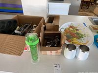 Cardboard boxes containing assorted knives, forks, spoons, a green plastic ice stick container, two white insulated Yeti-style glasses, and a metal tray with fruit motif
