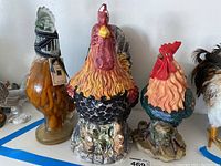 Five assorted rooster figurines including ceramic and glass materials, some with scratches and hand-painted details.