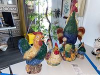Photo of assorted ceramic and glass rooster and chicken figurines arranged on a table in natural light. Includes larger roosters and smaller chickens with colorful detailing.