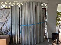 Mixed sizes of galvanized corrugated metal shutter panels leaning against the wall, secured with blue strap.