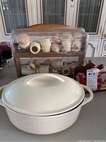 Overall view of the lot items including the wood spice rack wrapped in plastic with ceramic spice containers, white ceramic casserole dish with lid, and the wooden napkin holder with rooster decoration.