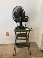 Cosco step stool with green seat and three metal steps, placed against wall with a black table fan on top.